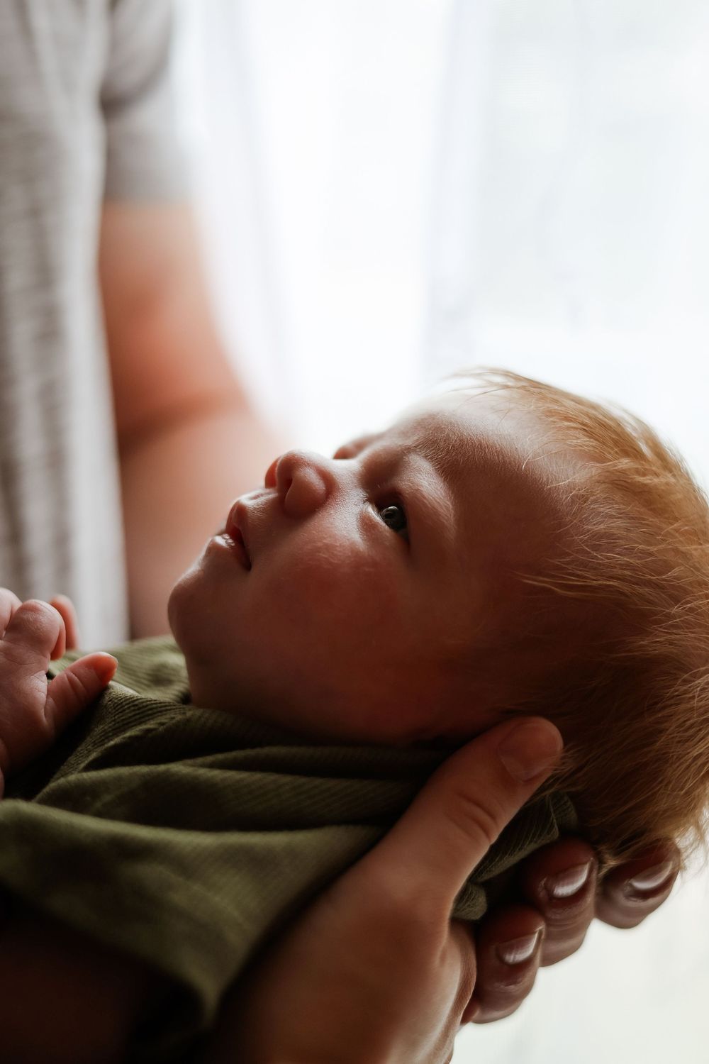 A newborn baby in a green onesie being cradled in adult hands, gazing upward in soft natural light.