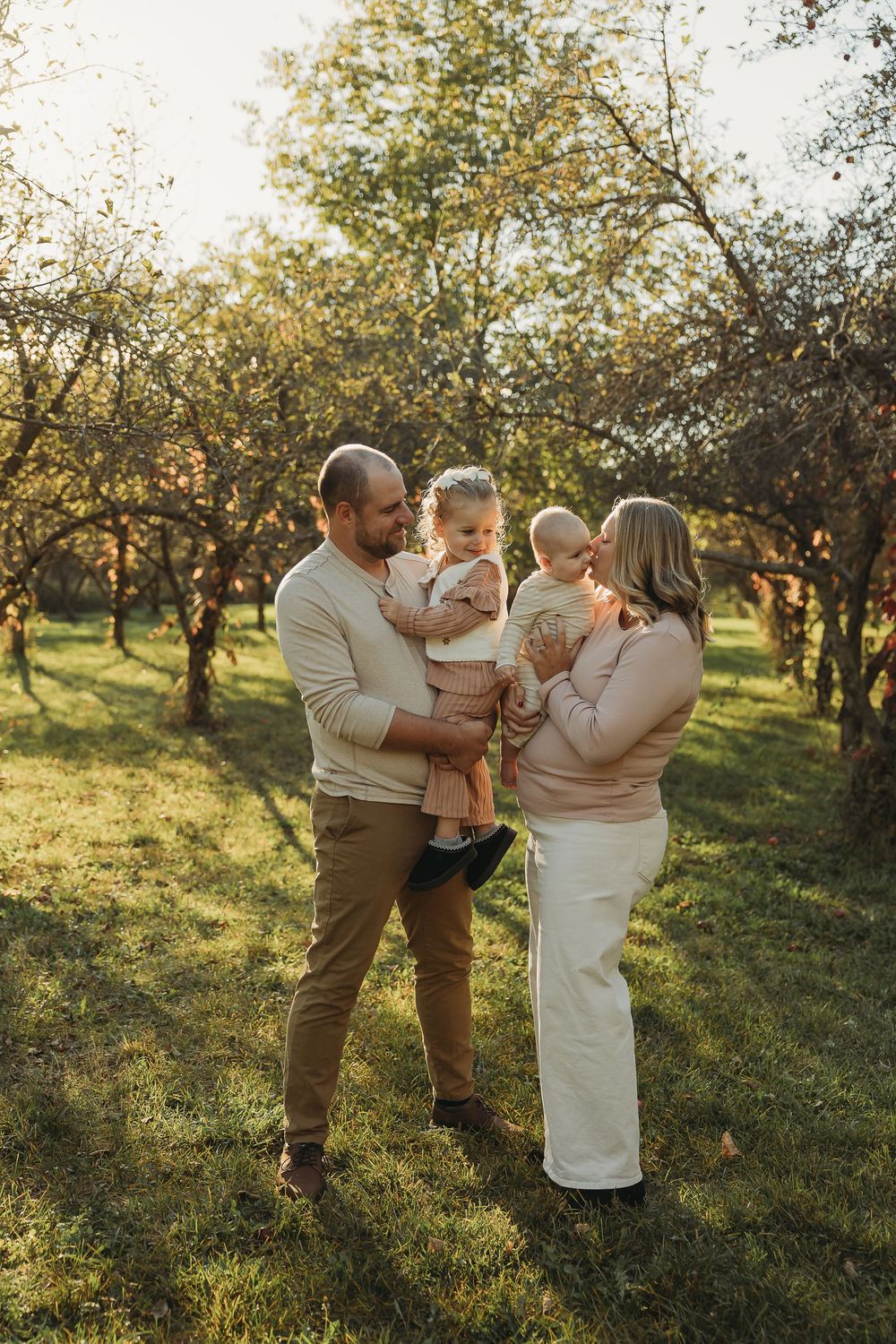 A family shares a tender moment together in an orchard at golden hour with sunlight filtering through the trees.