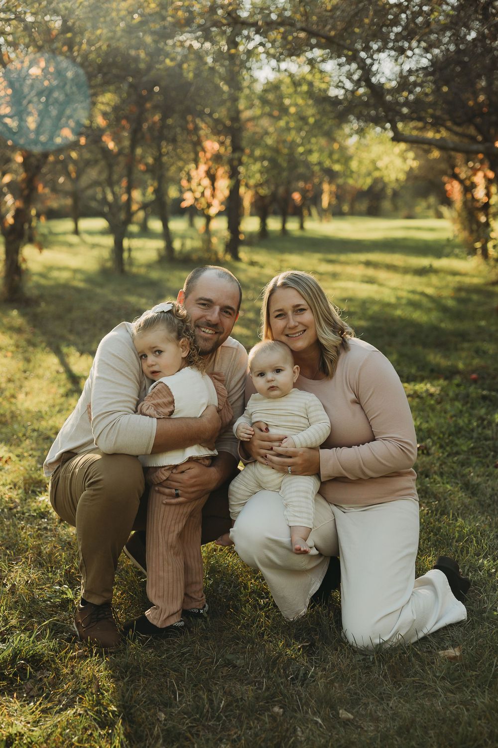 A family sits together on grass in an orchard during golden hour as sunlight filters through trees in the background.