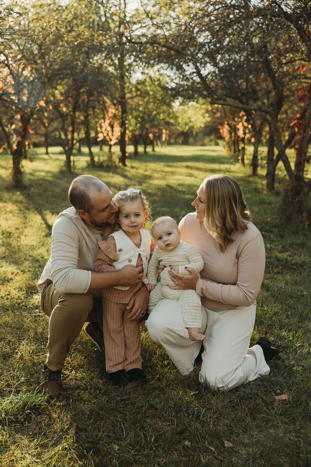 A family sits together in a sunlit orchard during golden hour wearing neutral toned clothing for a portrait session.