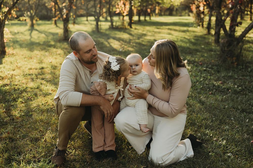 A family wearing neutral tones embraces in a sunlit wooded area during an outdoor photo session.
