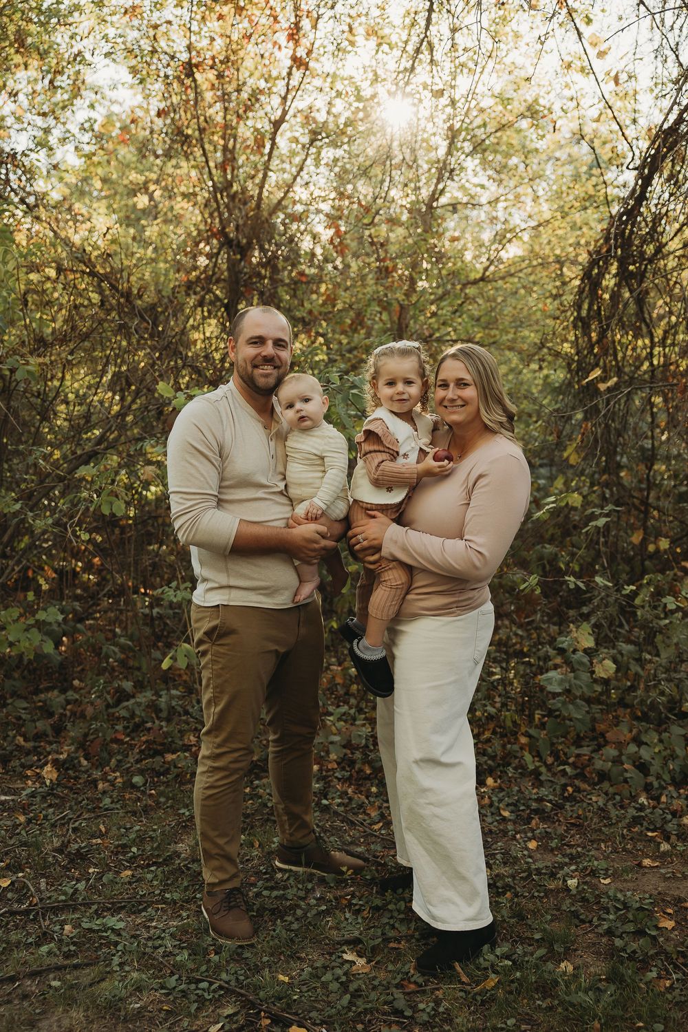 A family of four poses together in a wooded area during golden hour wearing neutral colored clothing.