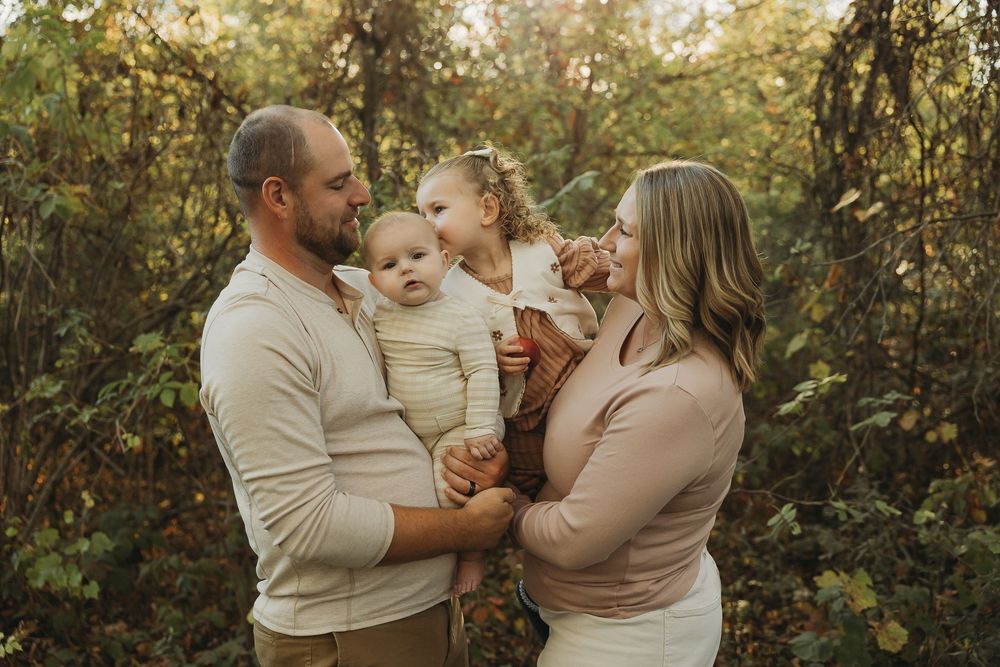 A family in neutral colored clothing shares tender moments together in a sunlit wooded setting during golden hour.