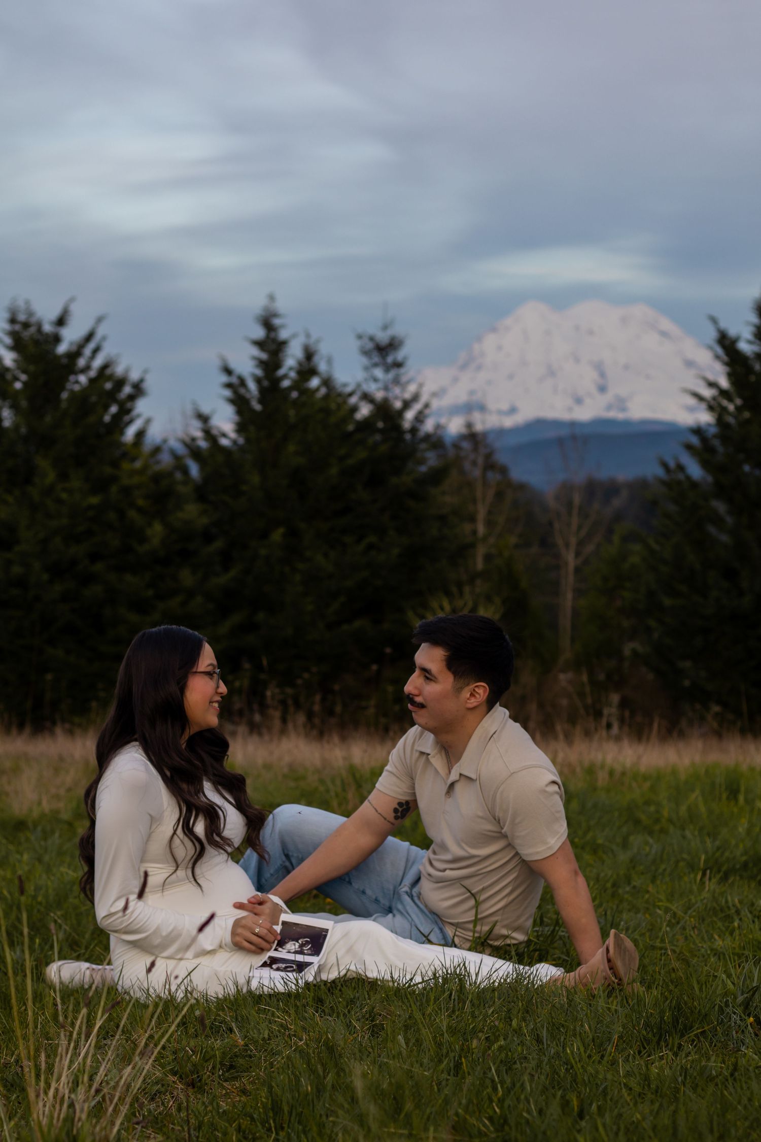 Couple sitting in grass field holding ultrasound photos with snow-capped Mount Rainier in background in Eatonville with Roy photographer