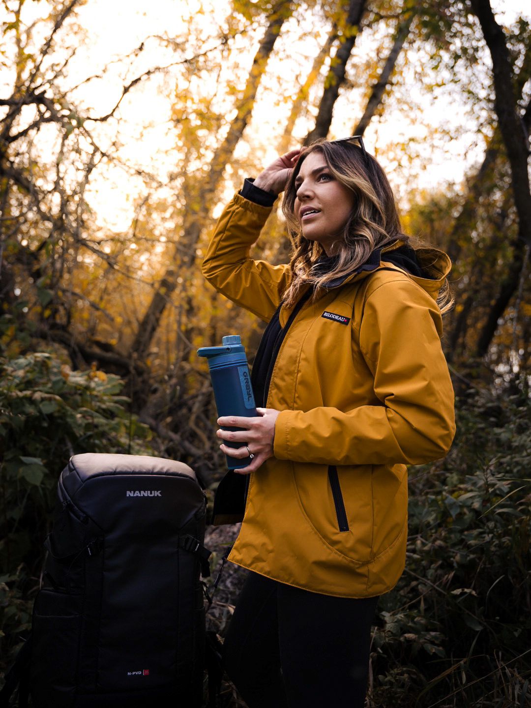 Person in yellow jacket holds blue travel mug while enjoying autumn forest scenery at sunset.