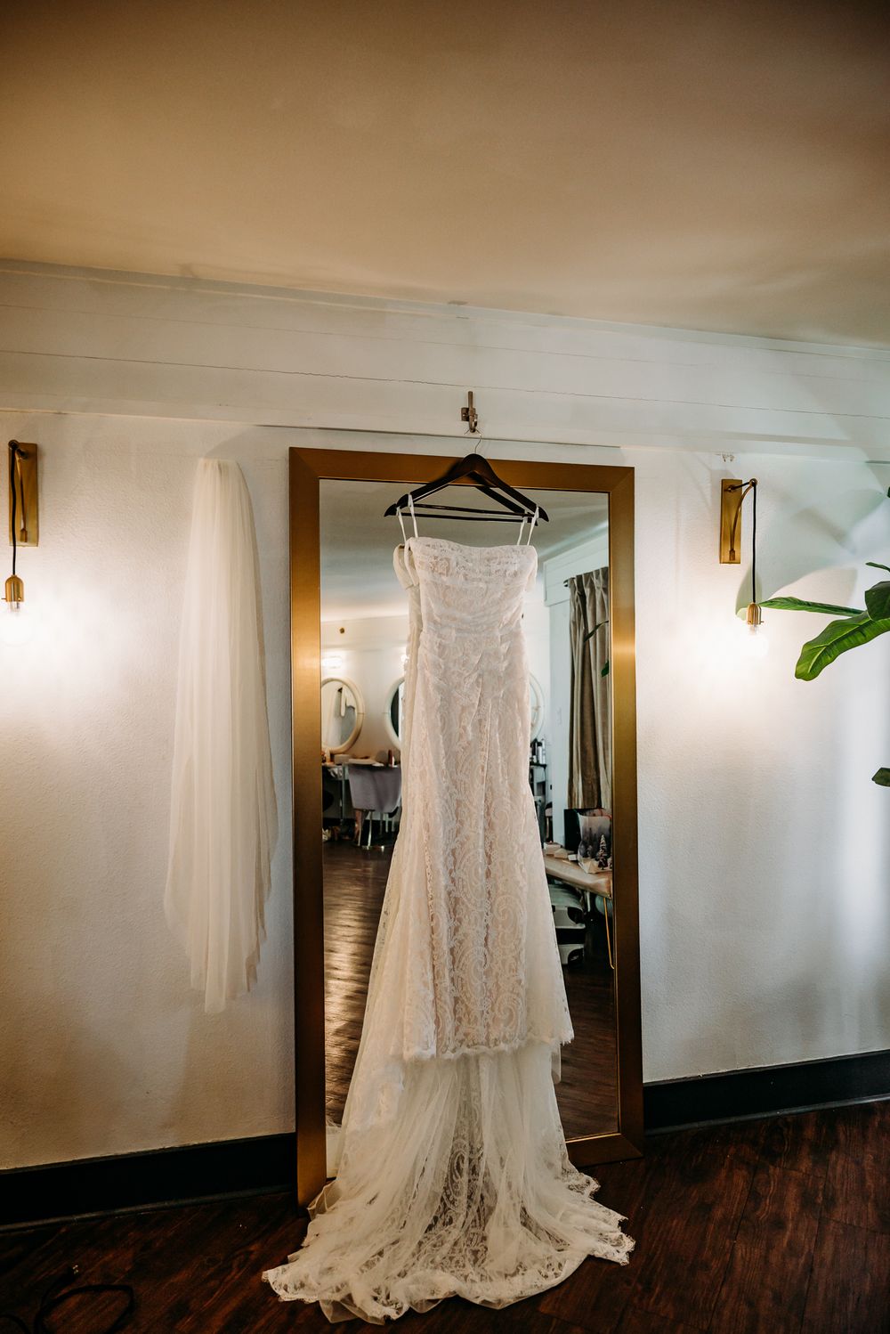 A beautiful lace wedding dress hangs gracefully against a white wall with warm lighting.