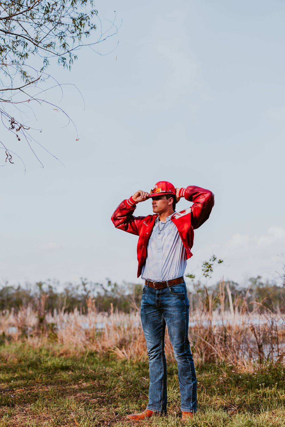 Person in red jacket and jeans stands in field against cloudy sky.