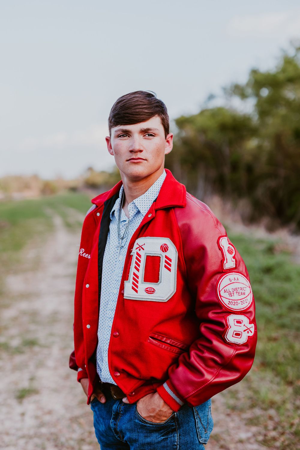 Person in red letterman jacket and blue jeans stands on rural path.