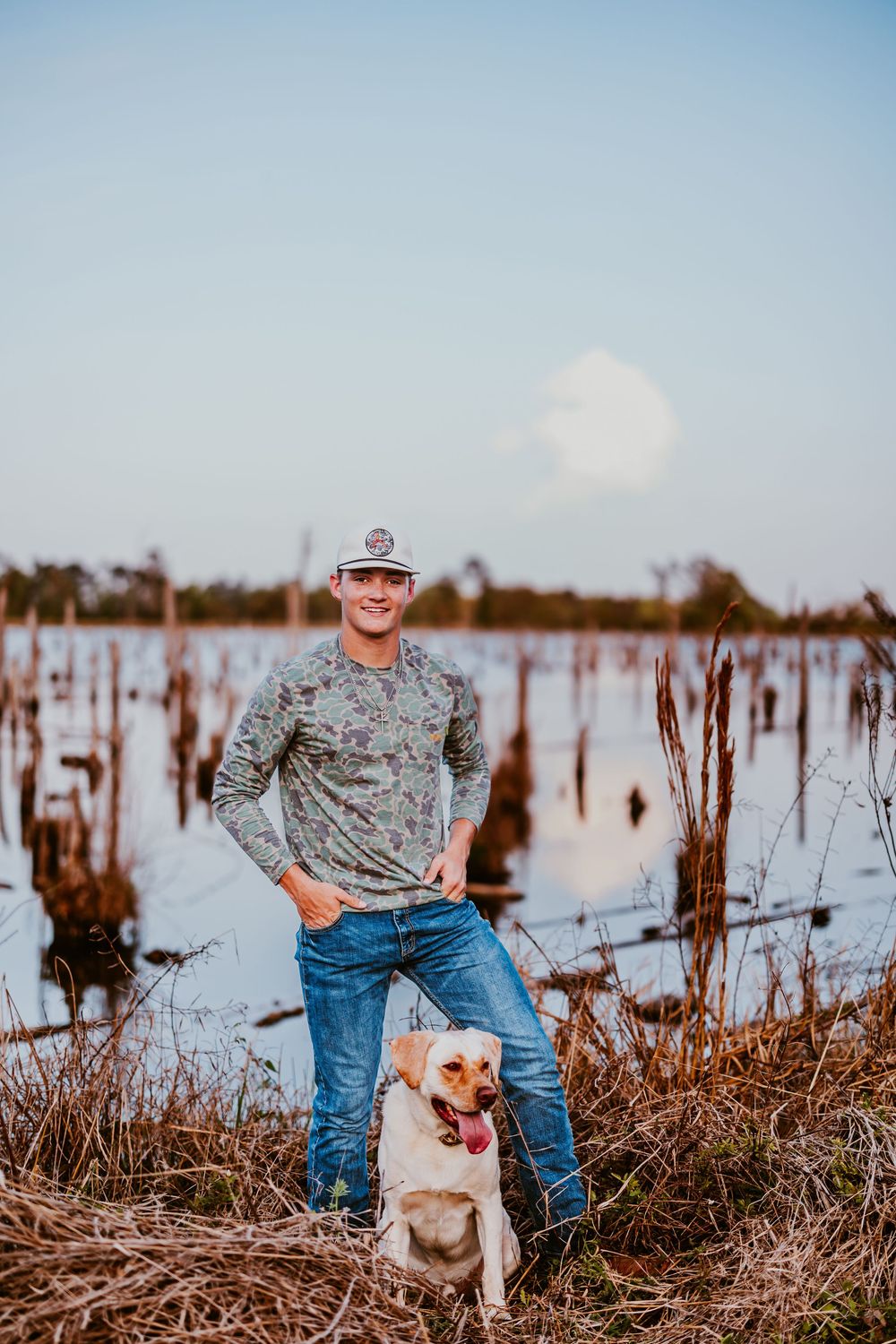 Person in camo shirt poses with small dog in marshy area.