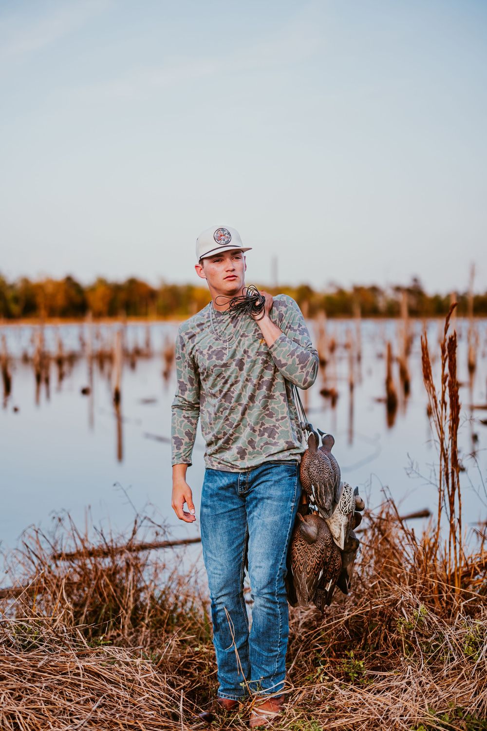 Person in camo shirt and cowboy hat stands among marsh vegetation.