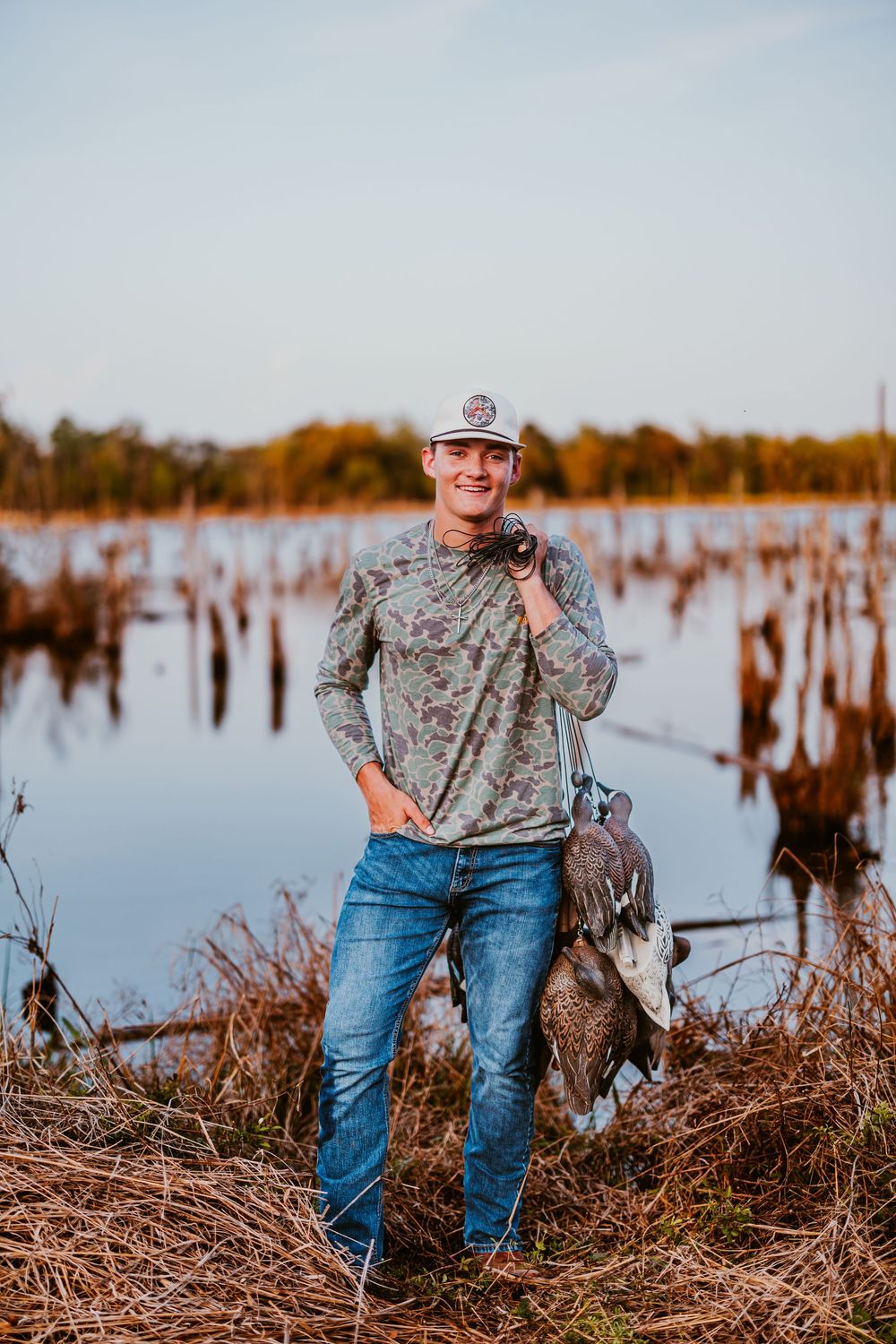 Duck hunter in camouflage gear stands near a marsh at sunset with hunting equipment.