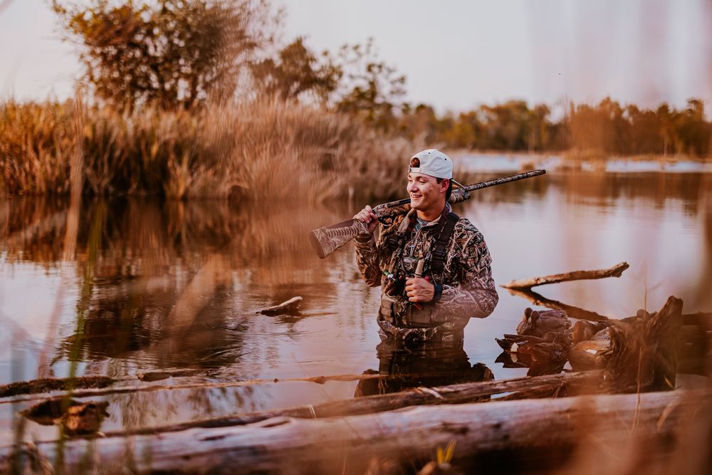 Waterfowl hunter in camo gear positioned in marsh during warm sunset light.