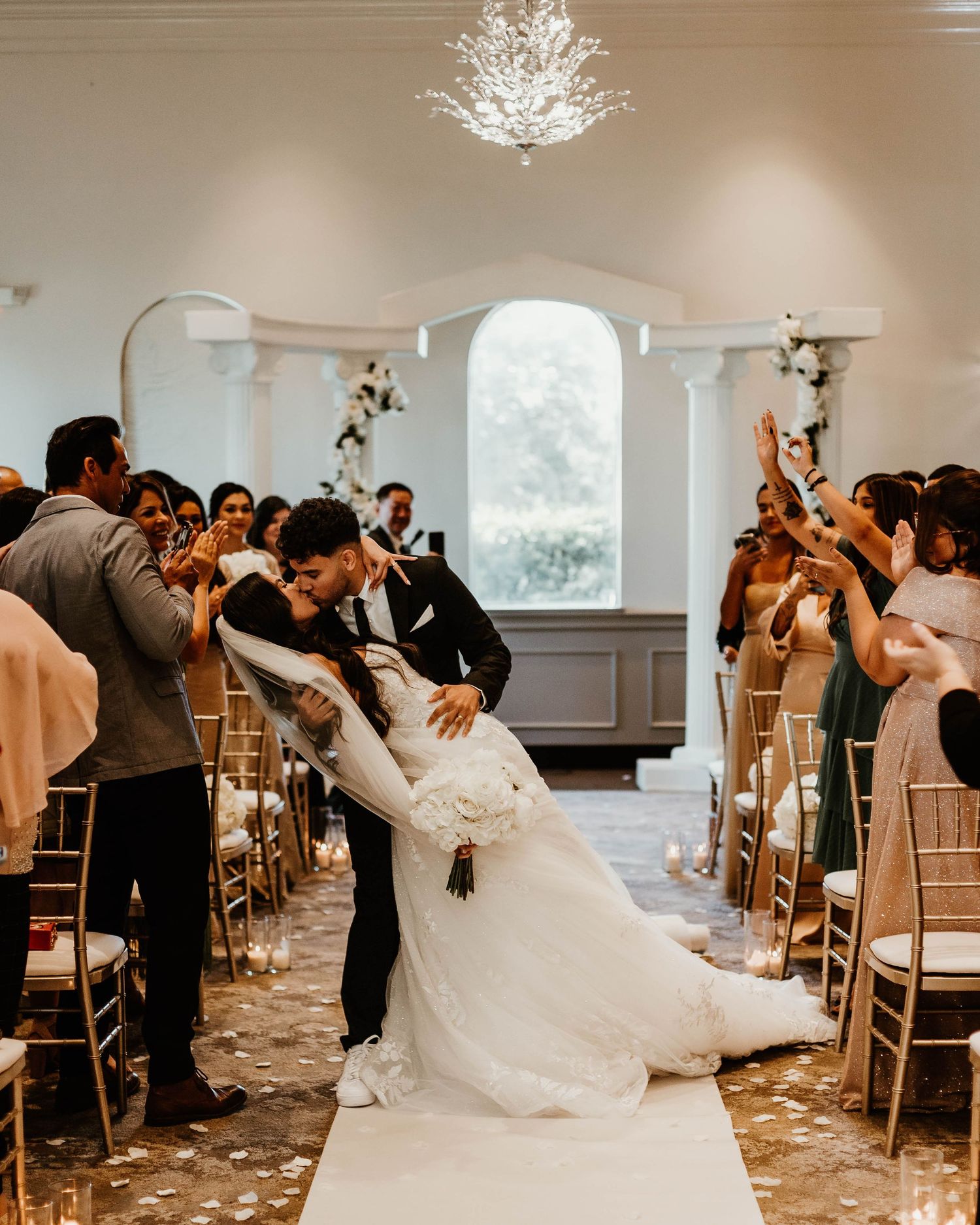 Wedding guests lean in for dramatic dip kiss during reception celebration with chandelier overhead.
