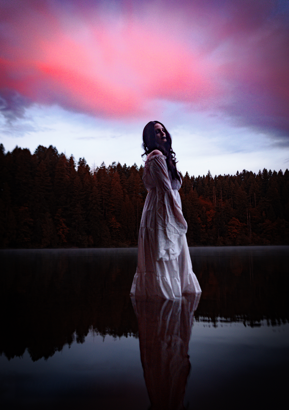 A silhouette in a white dress stands in still water at dusk with a dramatic pink and purple sky above forest treelines.