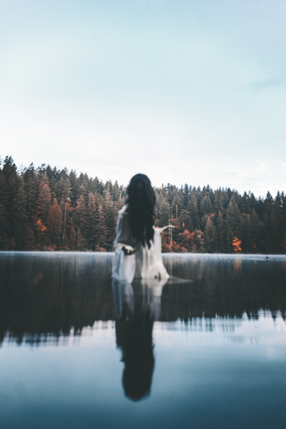 A dark silhouette stands in calm lake water with evergreen forest and autumn colors reflecting on the misty surface.