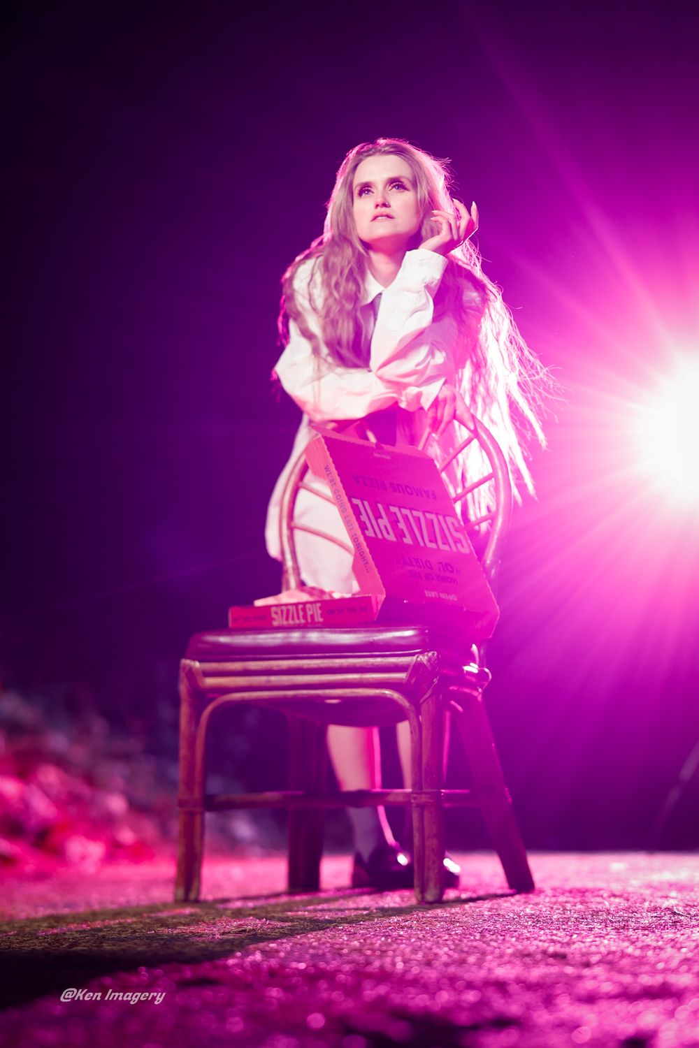A dramatic fashion photoshoot with a model seated on a chair against a vibrant purple and pink backlit background.