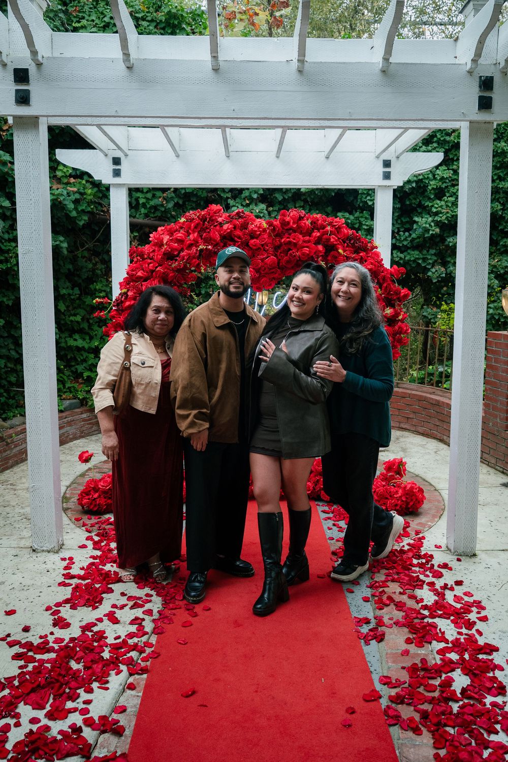Friends pose together under a white pergola adorned with bright red flowers and a red carpet.