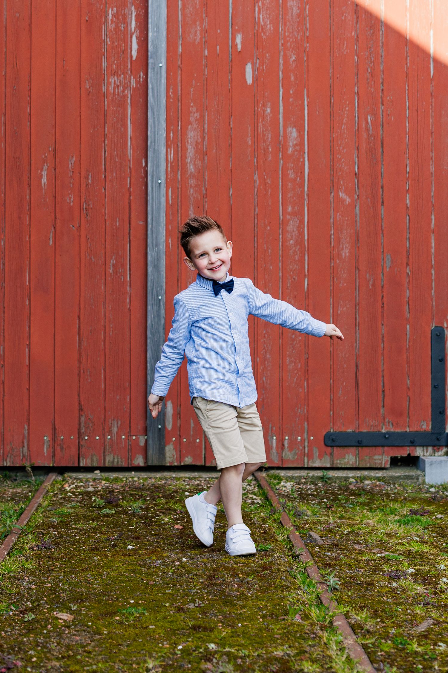 A child in a blue shirt and khaki pants playfully poses against a red barn wall in three connected photos.