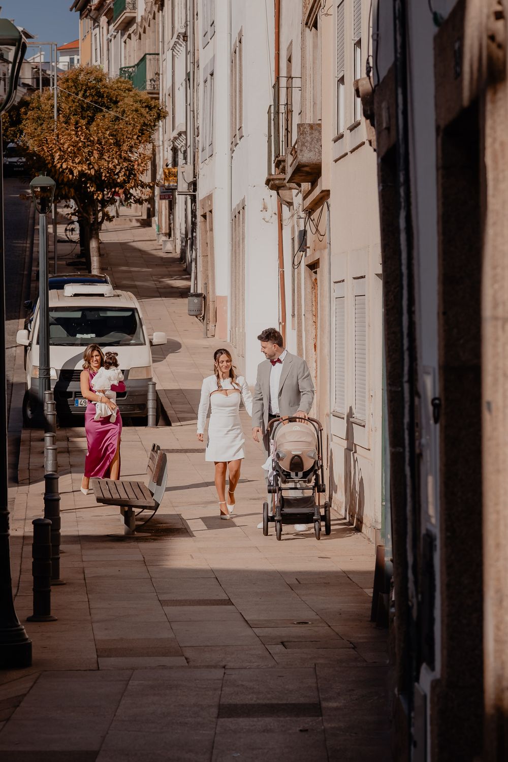 People walking on a sunny sidewalk with white buildings and parked cars along a European-style street.