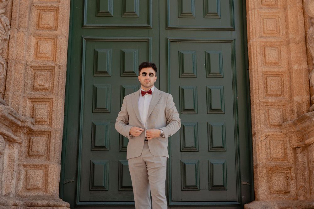A person in a light gray suit and red bowtie poses in front of large green wooden doors and stone archways.