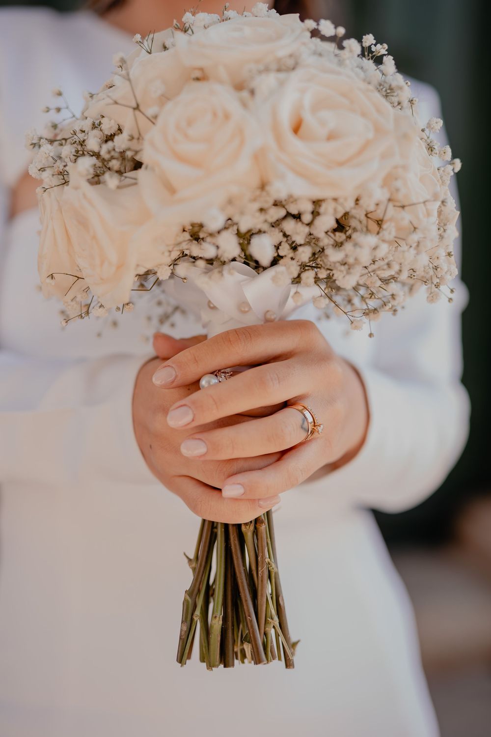 A delicate bridal bouquet of white roses and baby's breath held by hands with a sparkling engagement ring and manicured nails.
