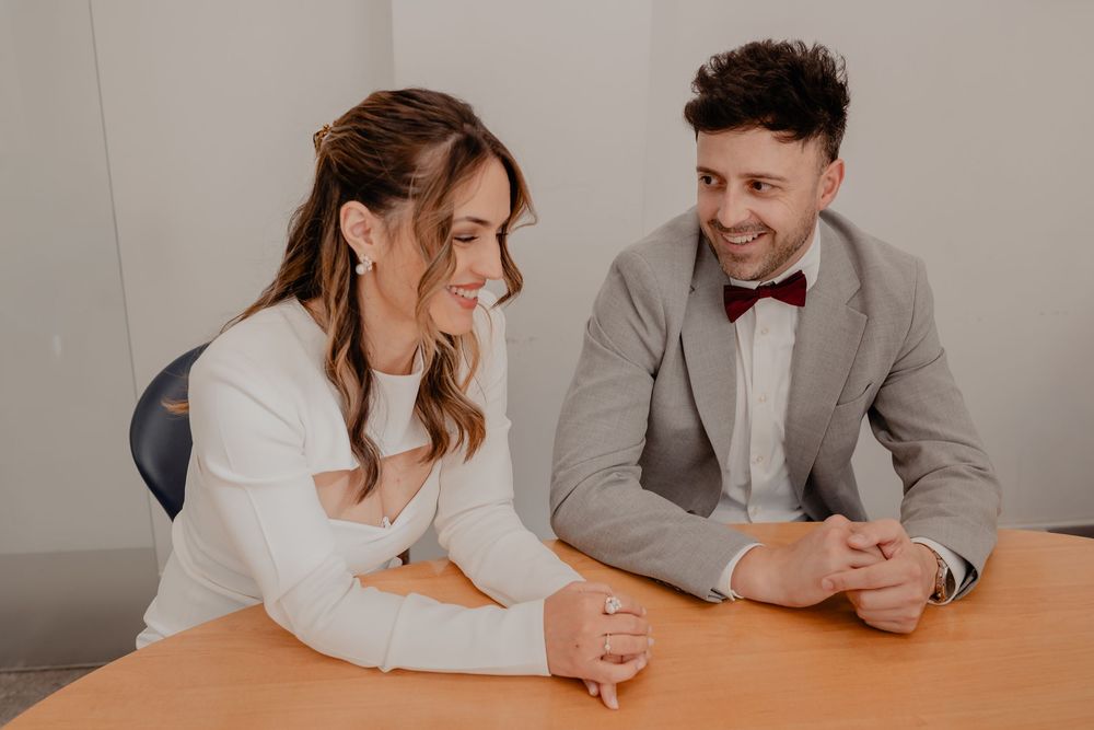A couple in formal attire sits at a wooden table sharing joyful moments together during a photoshoot.