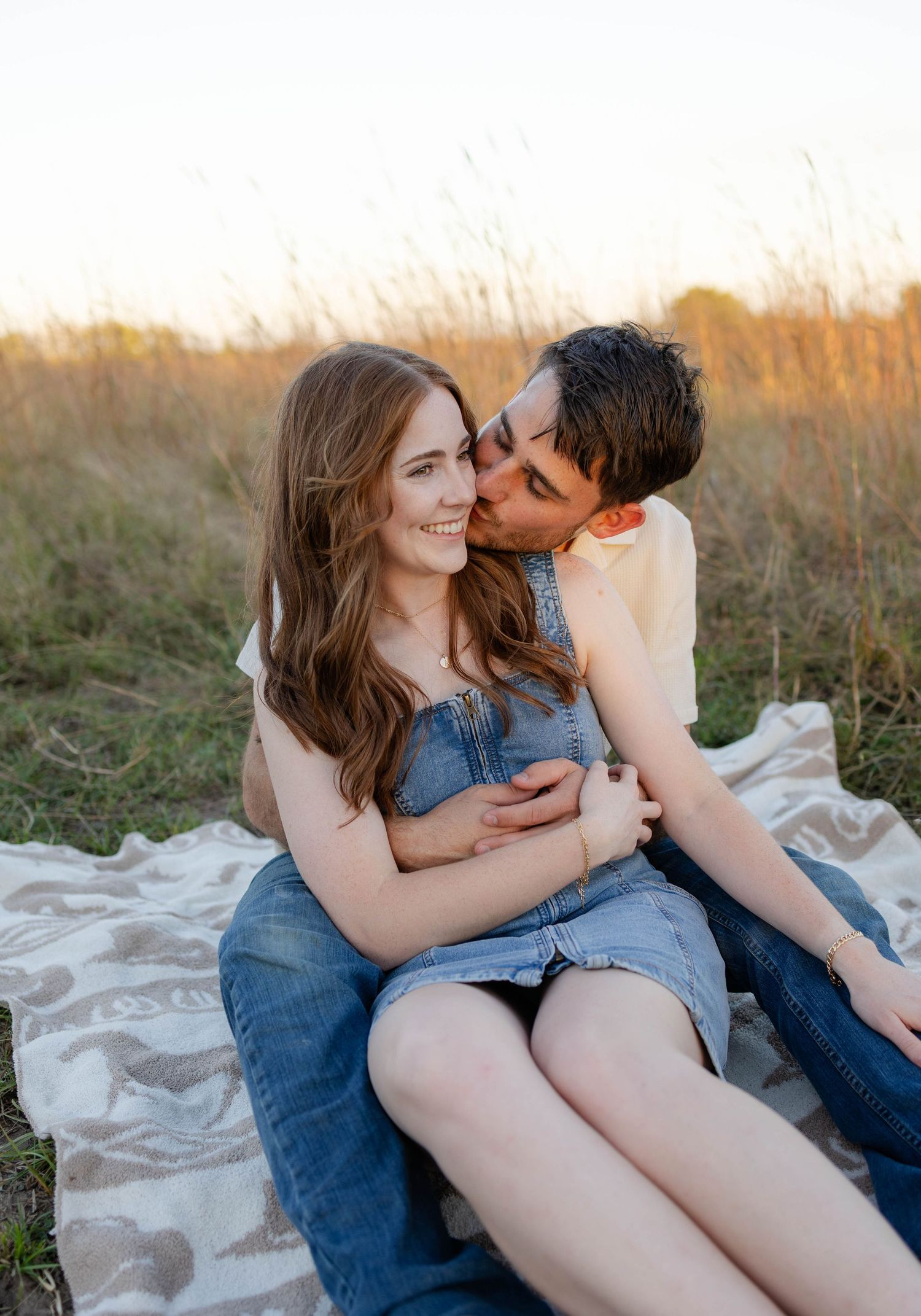 A couple shares an intimate moment while sitting on a blanket in a field during golden hour sunset.