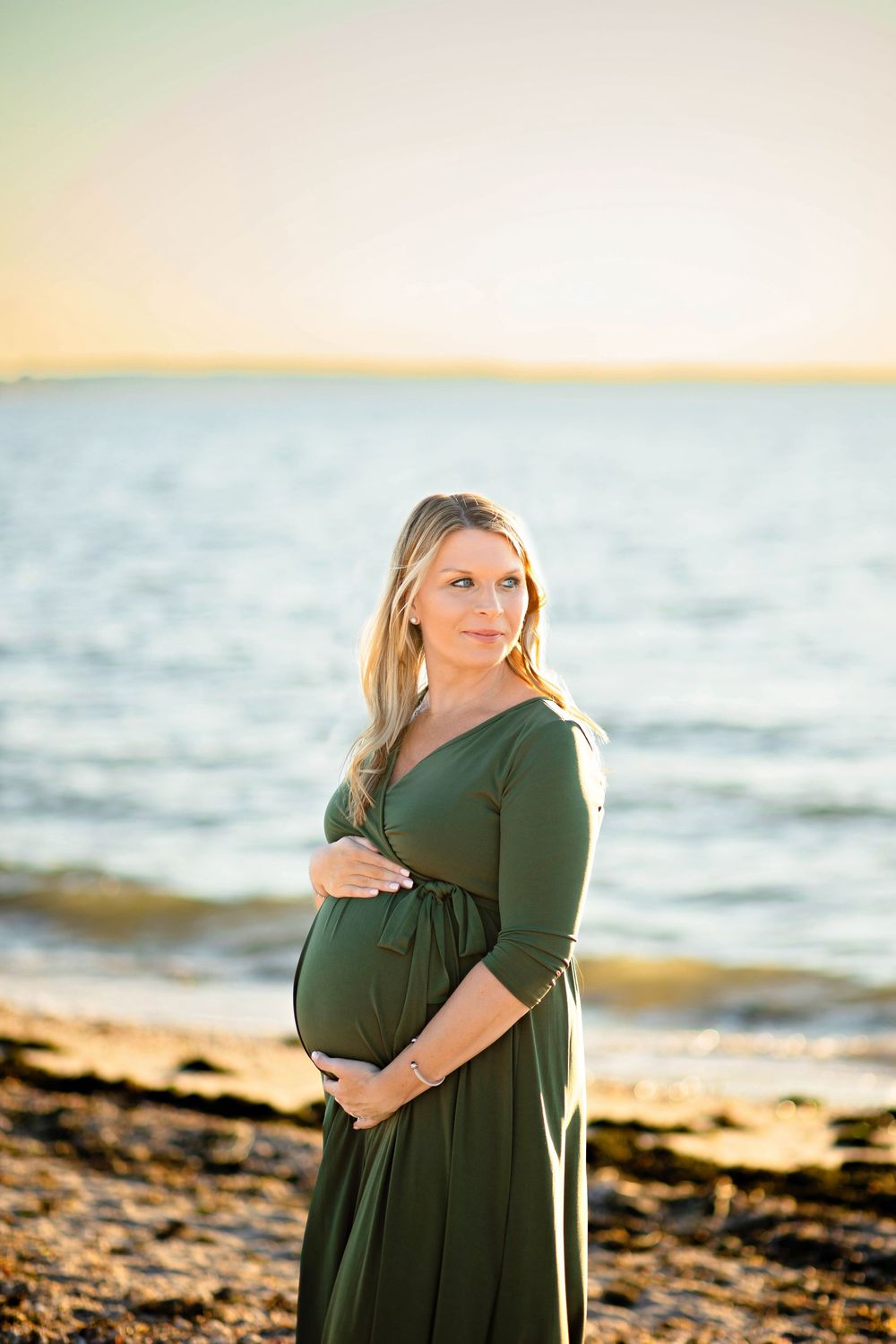 A pregnant woman in a green dress stands on a beach at sunset with gentle waves in the background.