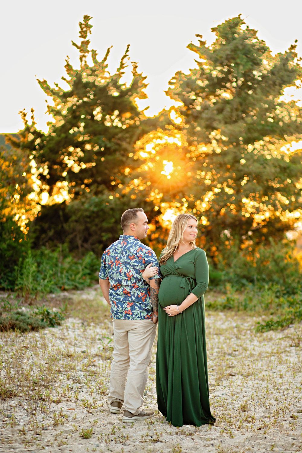 A couple shares a romantic moment in a field at golden hour as sunlight filters through evergreen trees in the background.