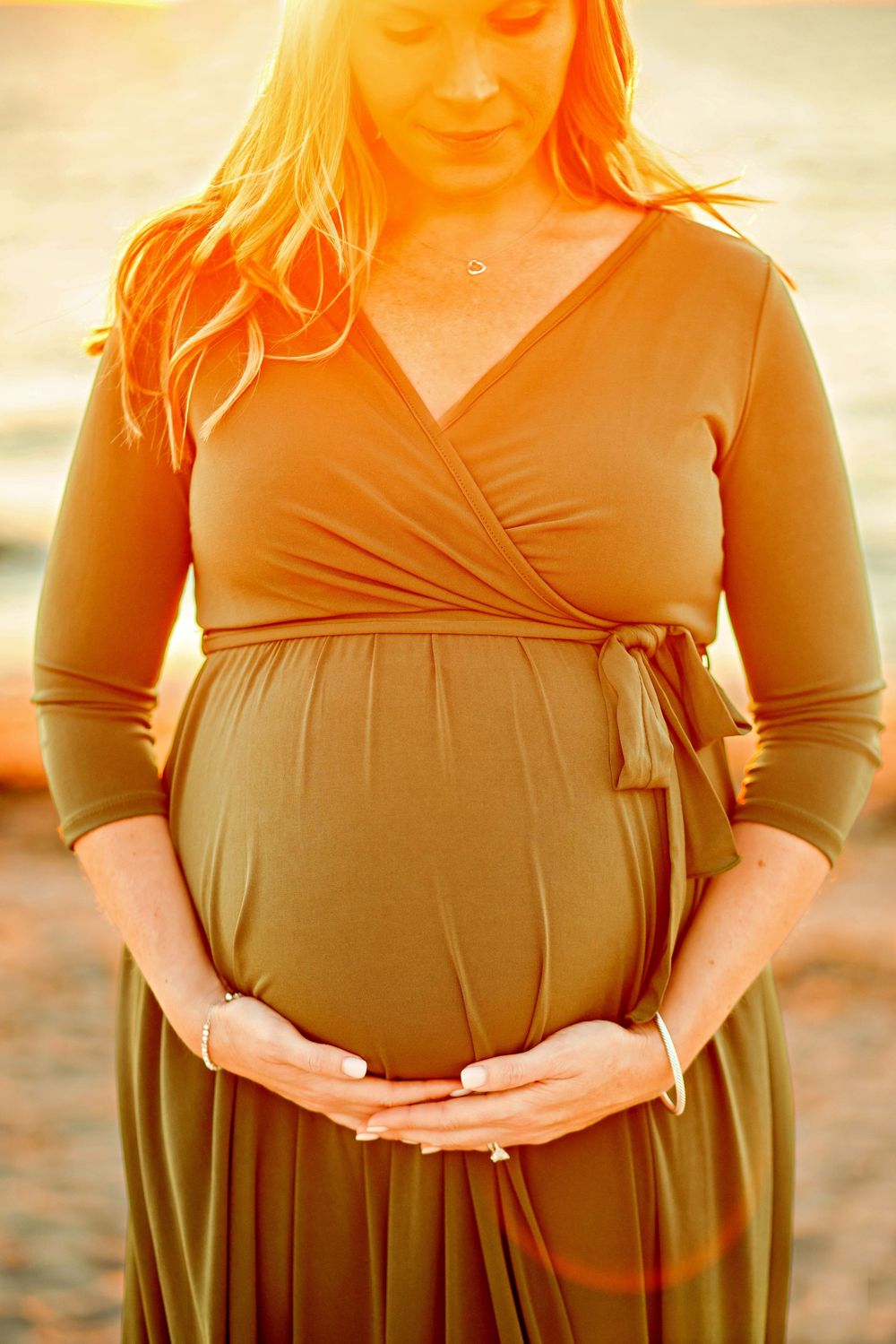 Pregnant woman in brown wrap dress holds her belly during golden hour sunset maternity photoshoot on beach.