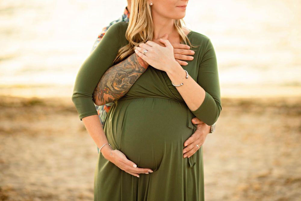 A person in an olive green maternity dress poses in a natural outdoor setting during a photoshoot.