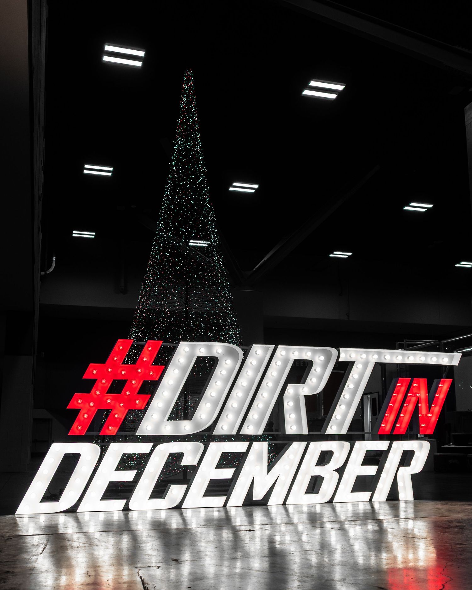 Illuminated Christmas tree silhouette with red and white #DIRT IN DECEMBER text display on reflective floor.