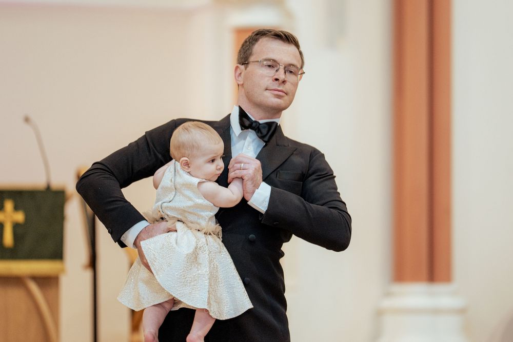 A baby in white baptismal clothing is lifted up during a christening ceremony at a church.