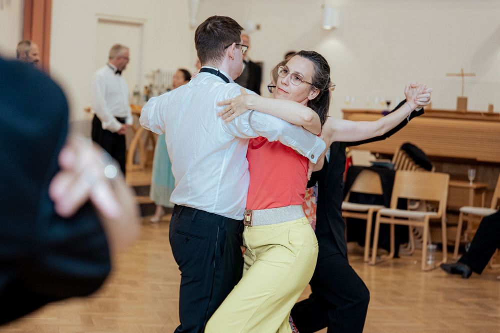 Dancers in bright casual attire embrace while practicing ballroom dance moves in a wooden-floored studio.