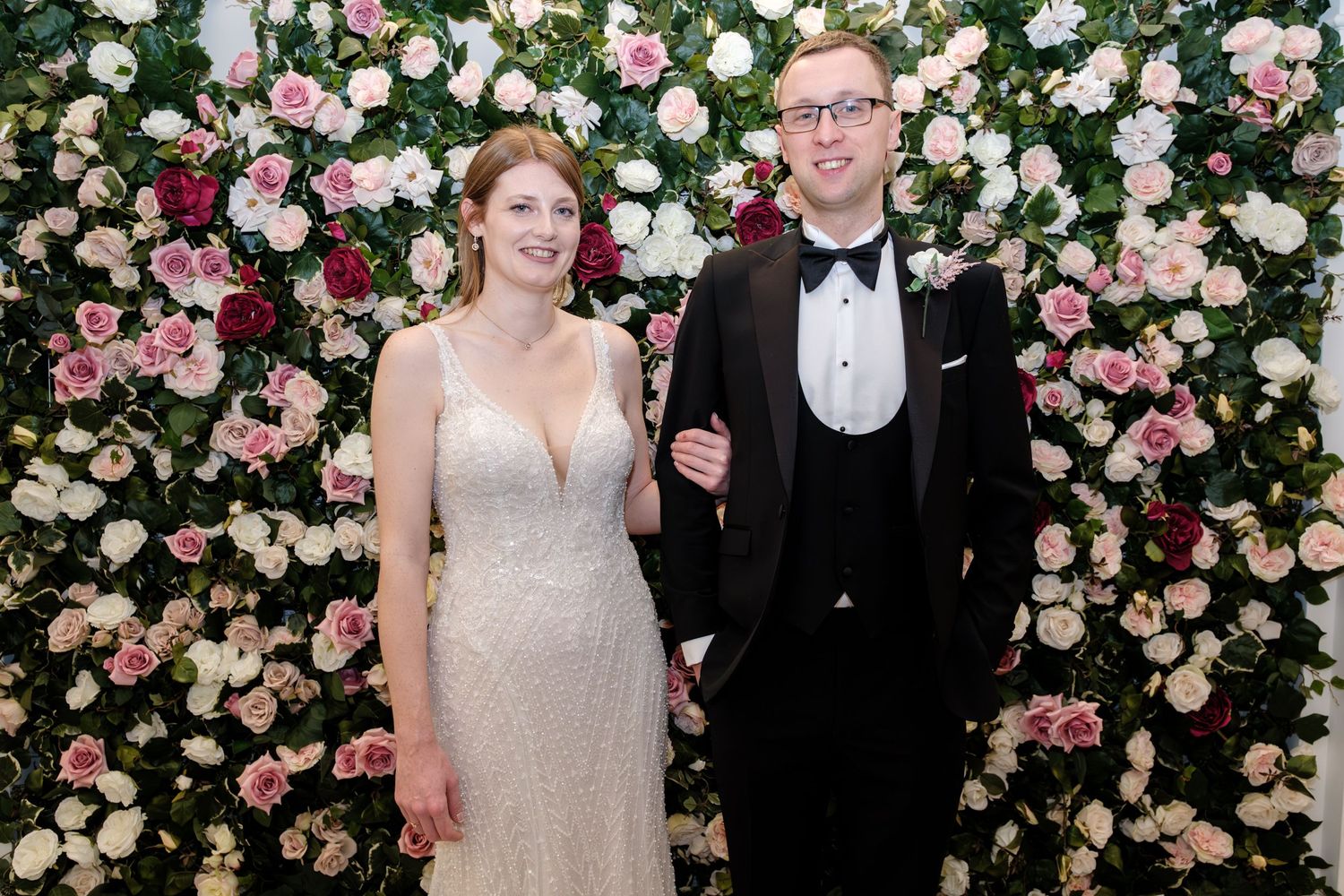A couple poses against a stunning wall of pink and white roses at an elegant wedding celebration.
