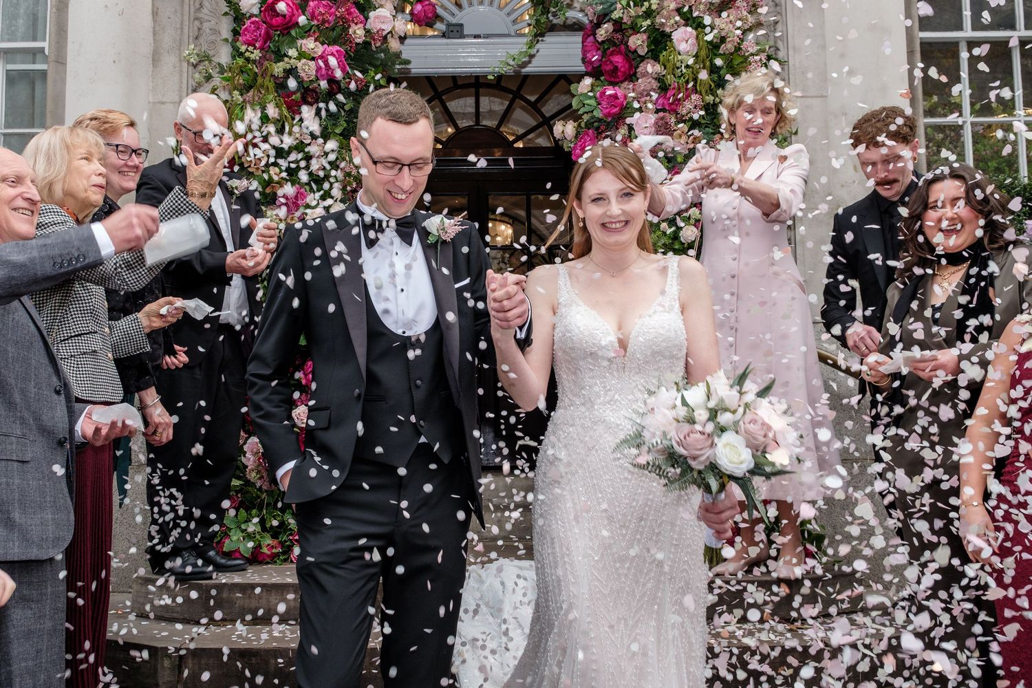 Wedding guests shower the newlyweds with pink rose petals during an outdoor celebration.