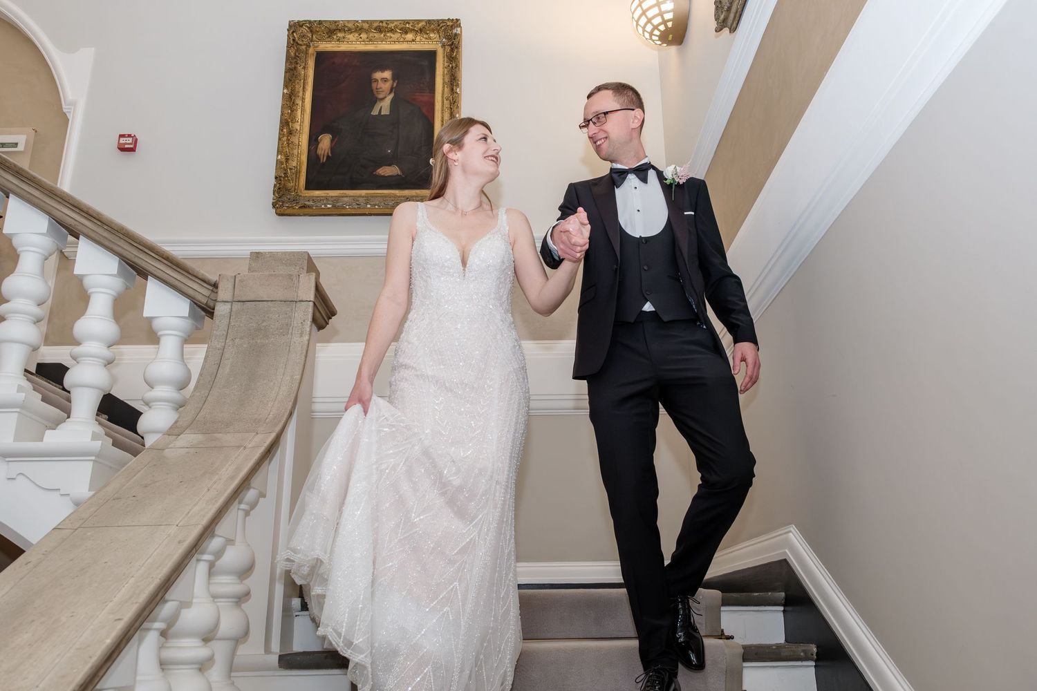 A couple in formal wedding attire poses on an elegant staircase with white walls and ornate gold-framed artwork.