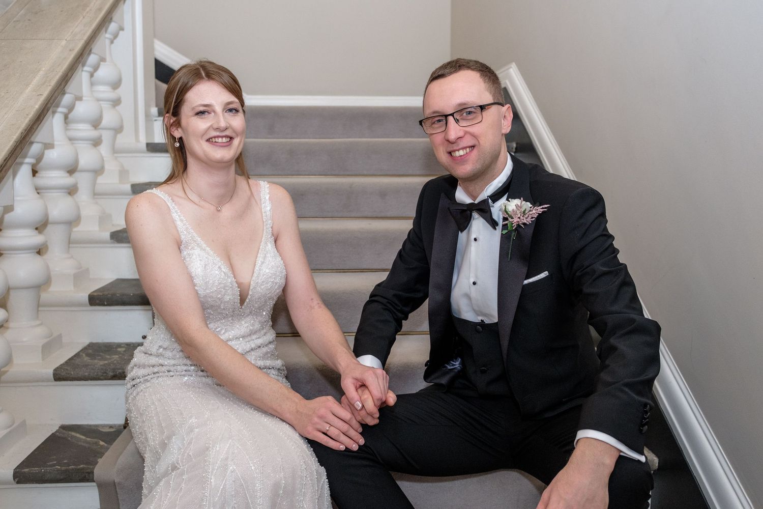 A couple in formal wedding attire sits together on elegant gray carpeted stairs.
