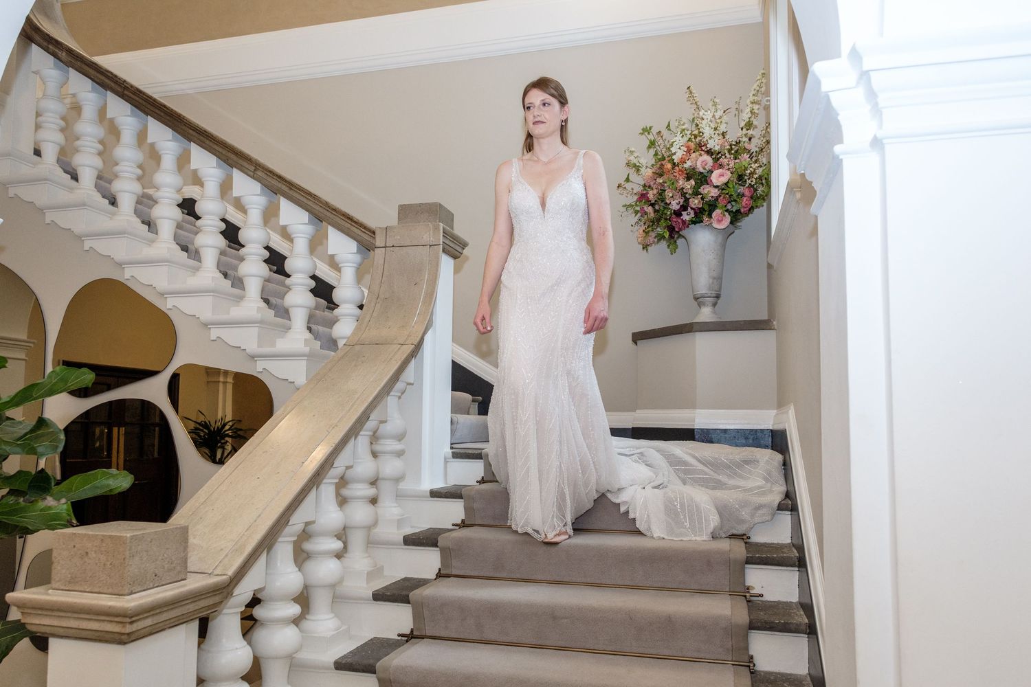 Elegant bride in flowing white gown poses gracefully on grand marble staircase in classic mansion setting.