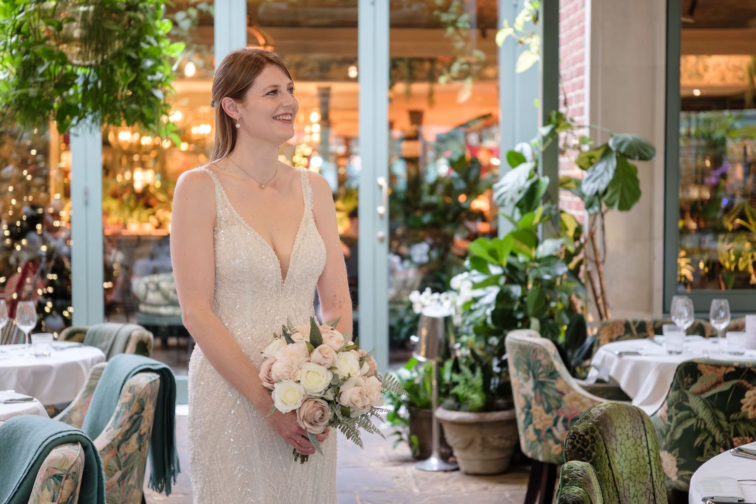 A bride in a white wedding dress holds a bouquet while standing in an elegant indoor venue with greenery and decor.