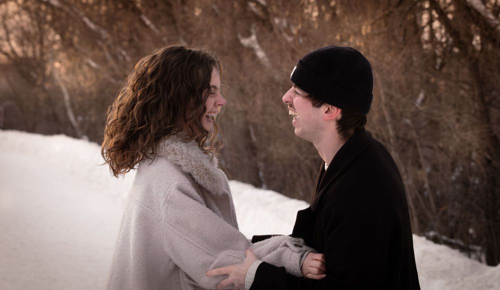 A couple shares an intimate moment while standing in snowy winter scenery with brown trees in the background.