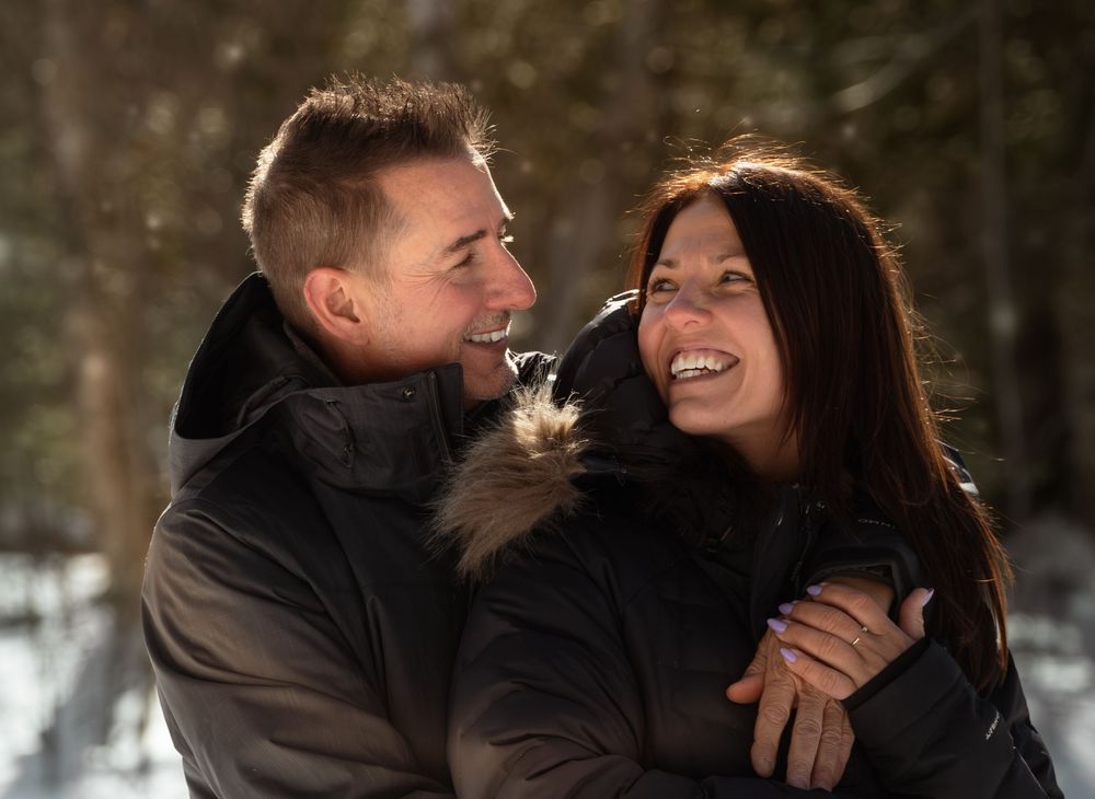 A couple laughs and embraces while enjoying a romantic moment in a snowy winter forest setting.