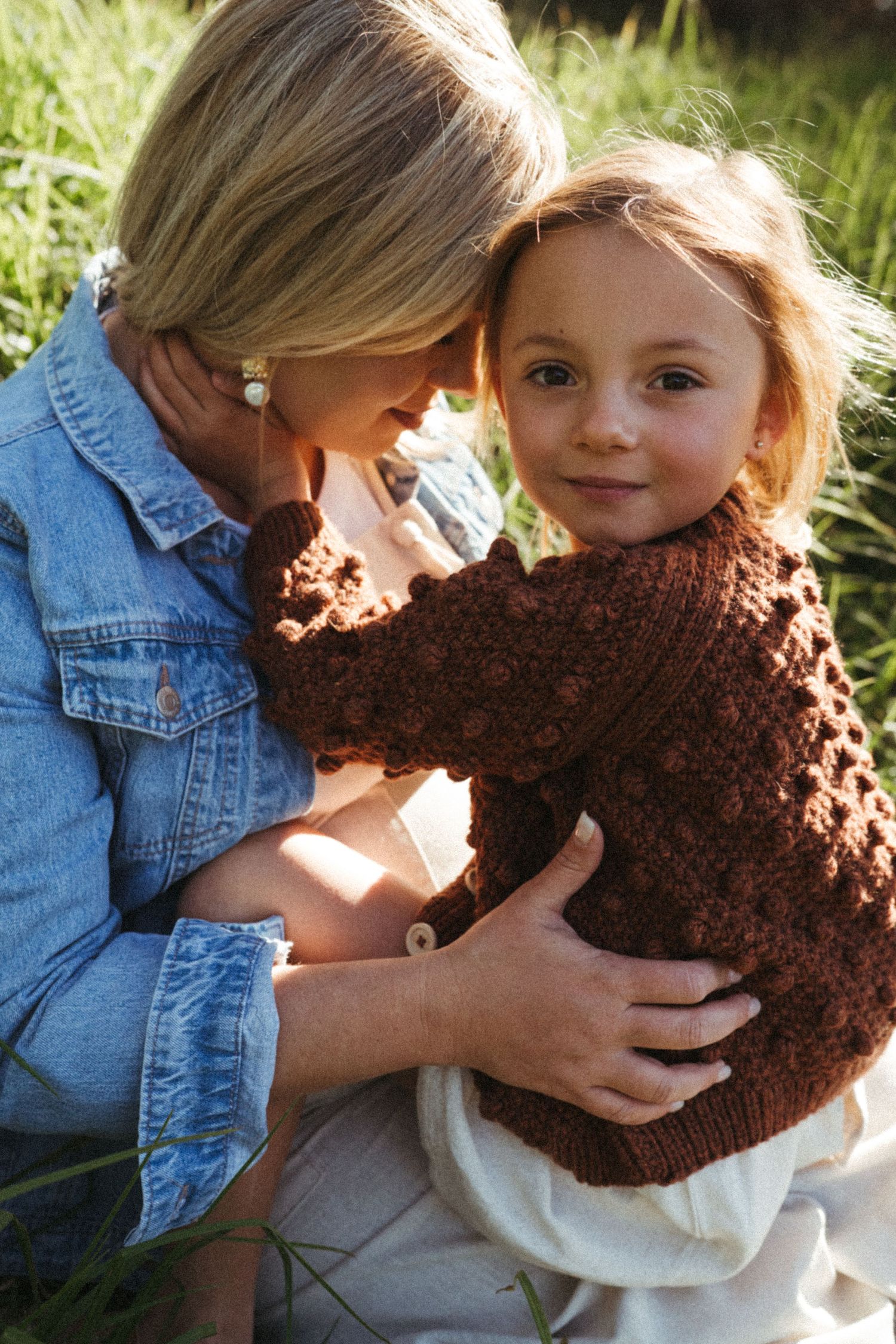 Two people lean close together outdoors while holding a brown knitted item.