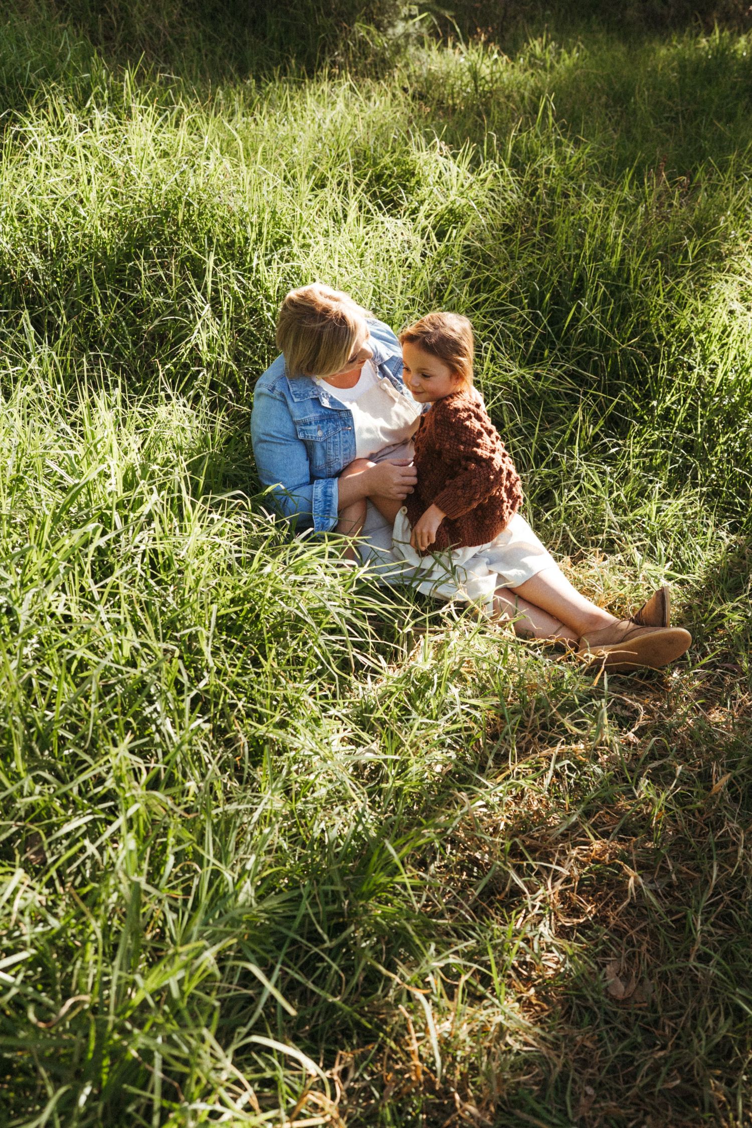 Two figures sitting together in tall sunlit grass wearing white and brown clothing.