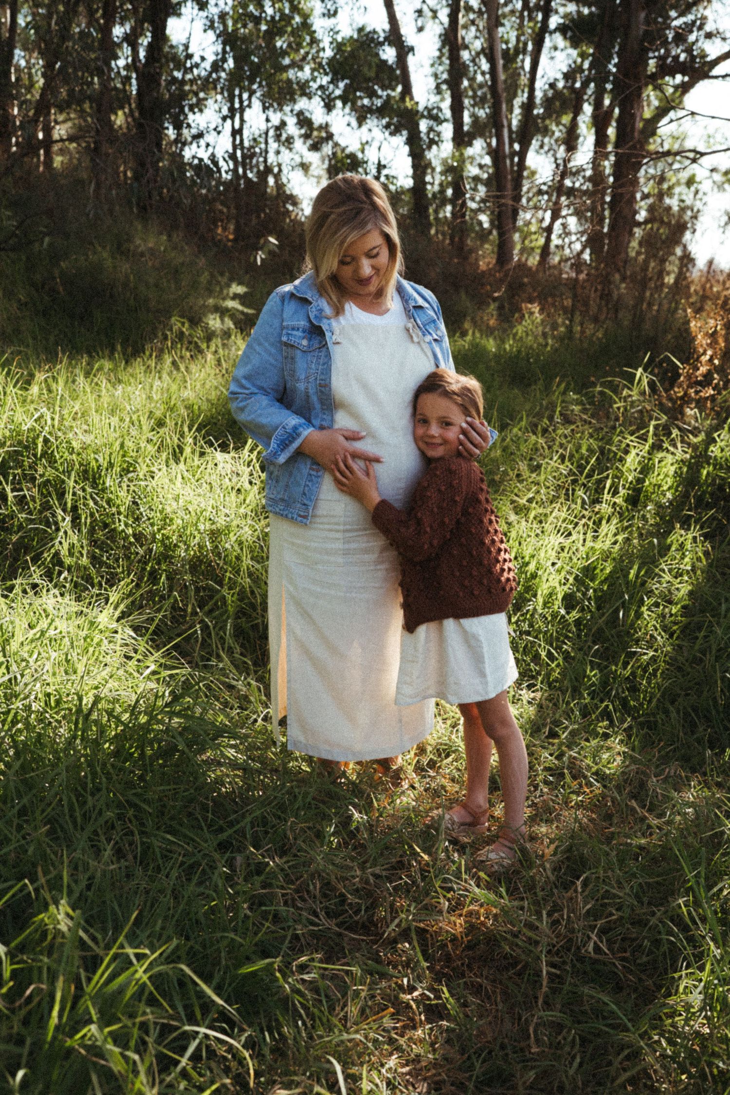 A tender outdoor family moment captured in sunlit grass with eucalyptus trees in background.
