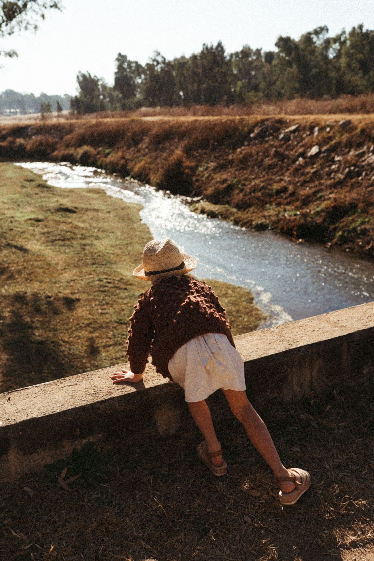 Figure in straw hat and brown sweater leaning over concrete wall near flowing stream.