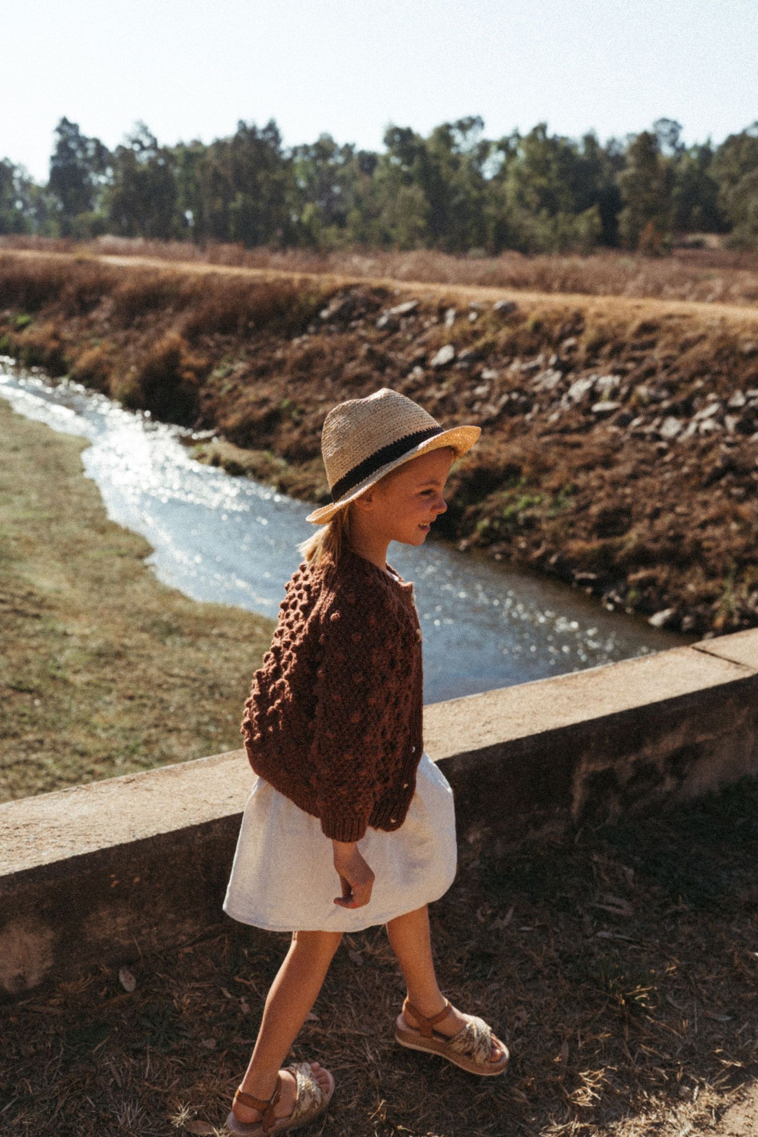 Person in brown sweater and straw hat walking along stream bank on sunny day.
