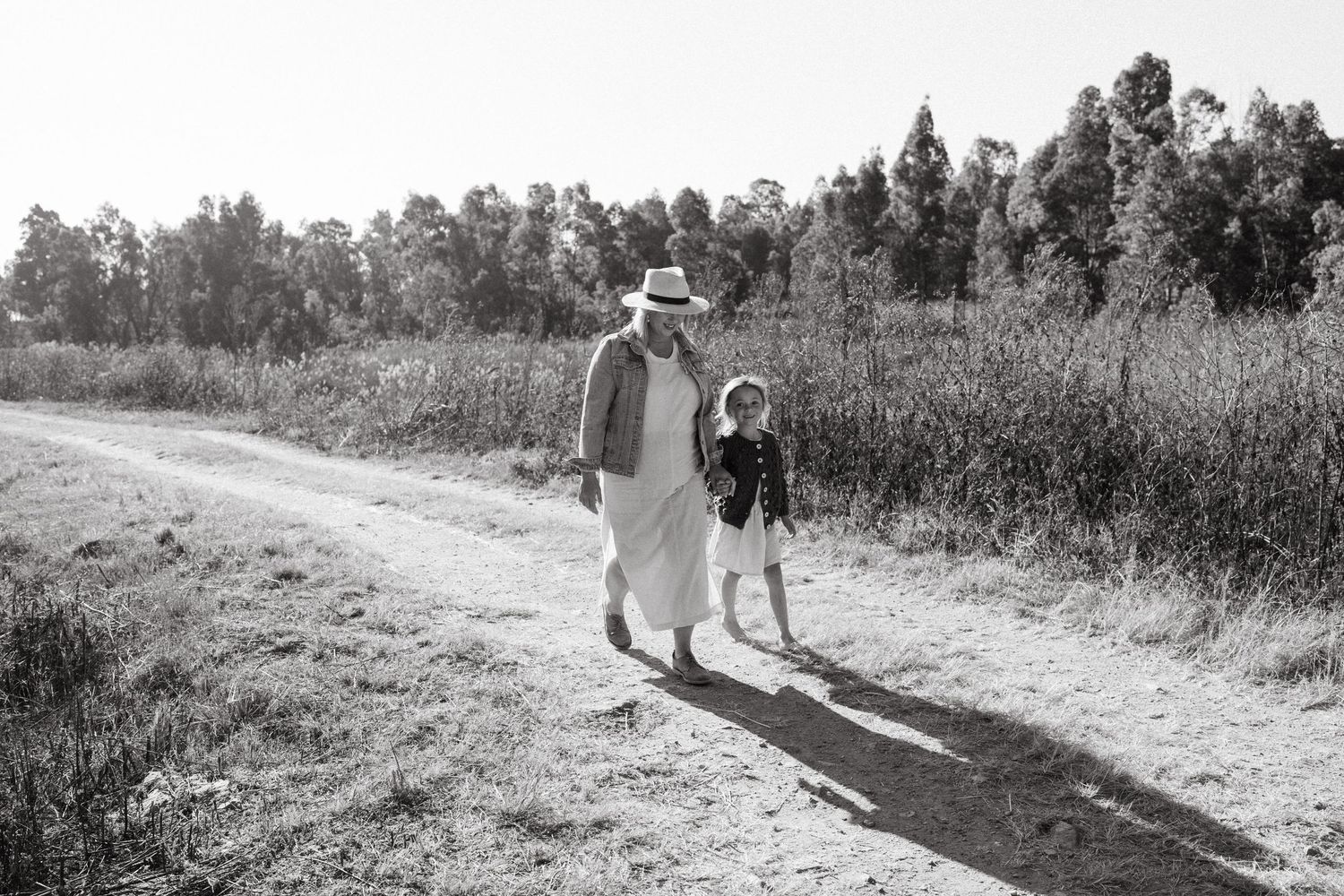 Black and white photo of two people walking along rural path with tall grass and trees.