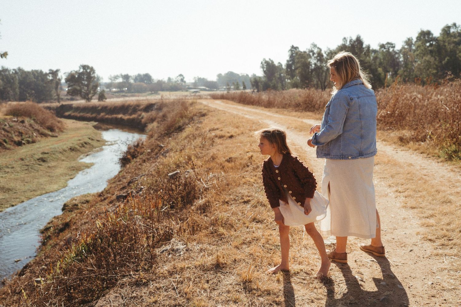 People walking along a dusty trail beside a stream on a sunny afternoon.
