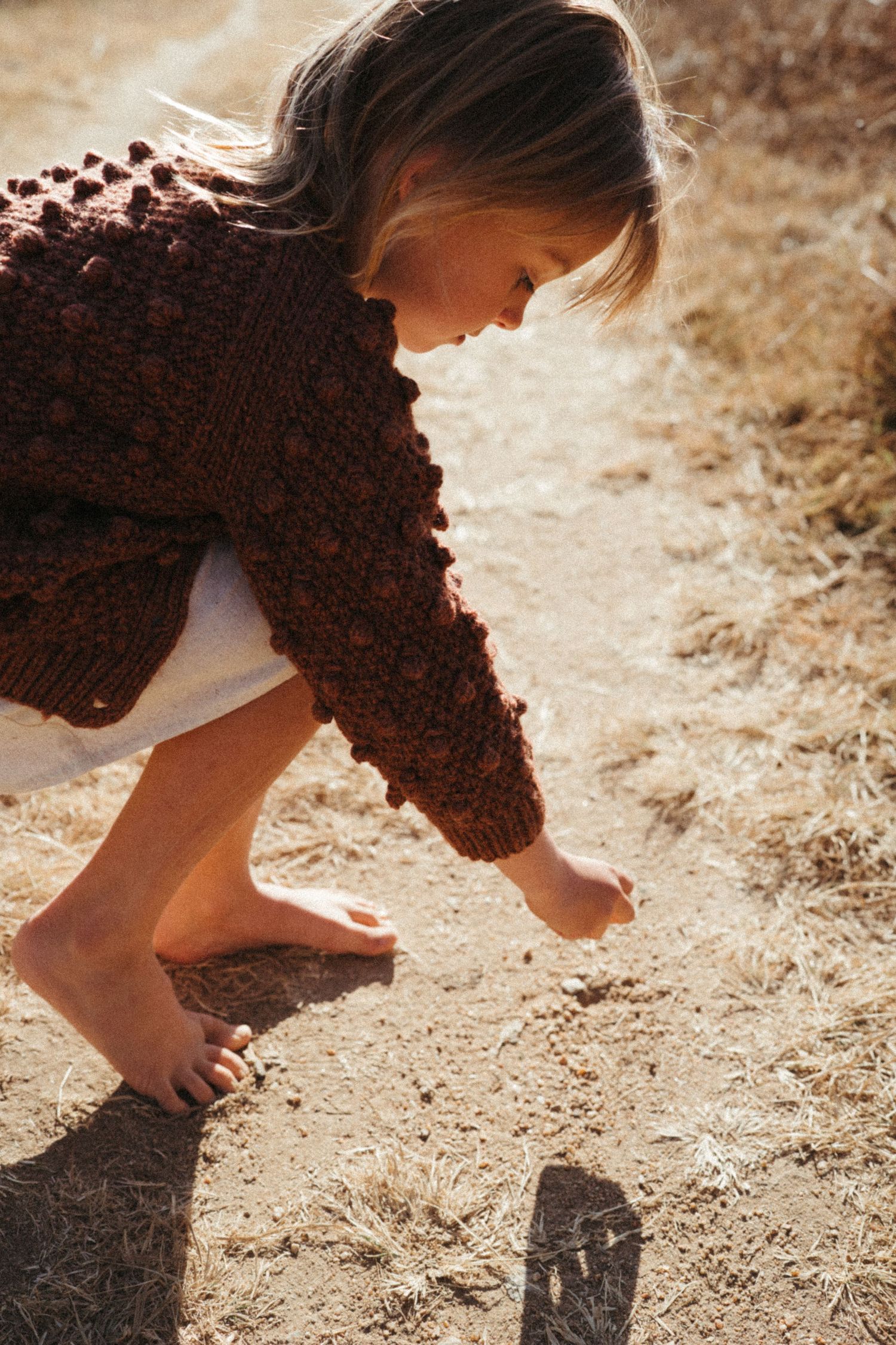 Bare feet touching the dusty ground in warm sunlight wearing a brown knit sweater.